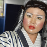 Close-up of a person in elaborate makeup and a striped kimono-style robe, wearing a styled wig with black and white hair.