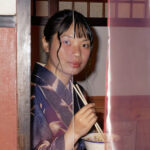 Woman in kimono eating ramen, partially obscured by a curtain.