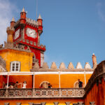 Pena Palace in Portugal, featuring a vibrant yellow and red exterior, clock tower, and people on a balcony.